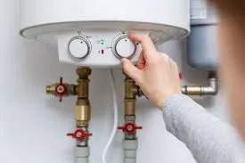 A person adjusts the temperature dial on a white water heater with visible pipes and red valve handles below.